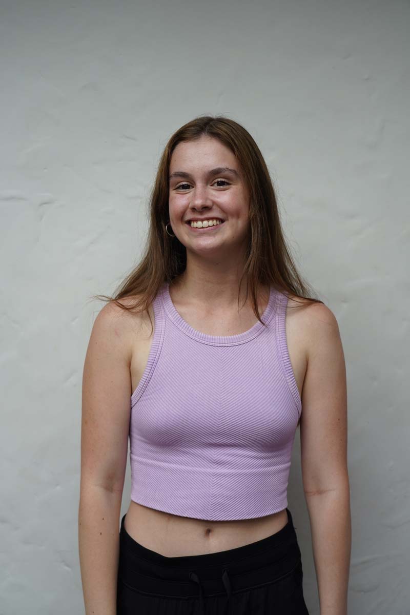 A person with long dark blond hair and a lavender tank top smiles at the viewer.