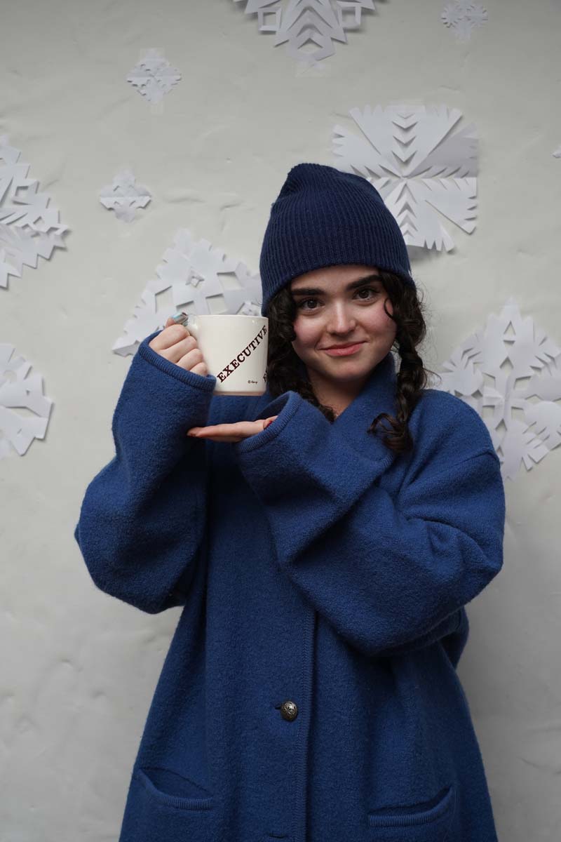 A person with a dark blue winter cap and a heavy dark blue winter coat stands in front of a wall of paper snowflakes, smiling at the viewer and holding a ceramic mug with the word "Executive" on it in an all-caps typeface. The typeface is Clarendon bold, and it is beautiful.