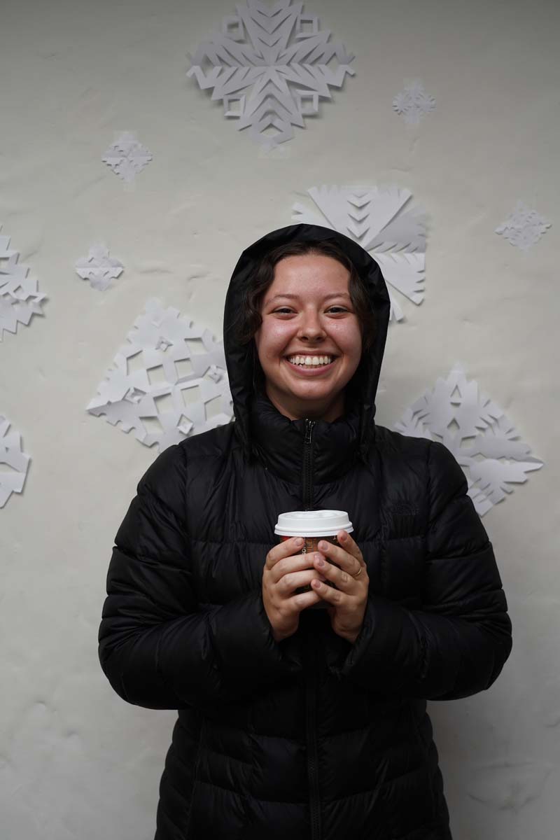A person wearing a black hoodie holds a coffee cup and smiles at the viewer.