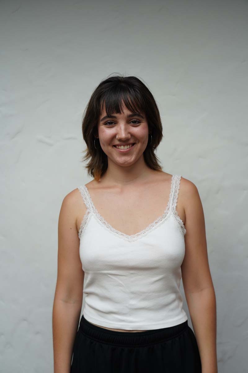 A person with long brown hair and a white tank top smiles at the viewer.