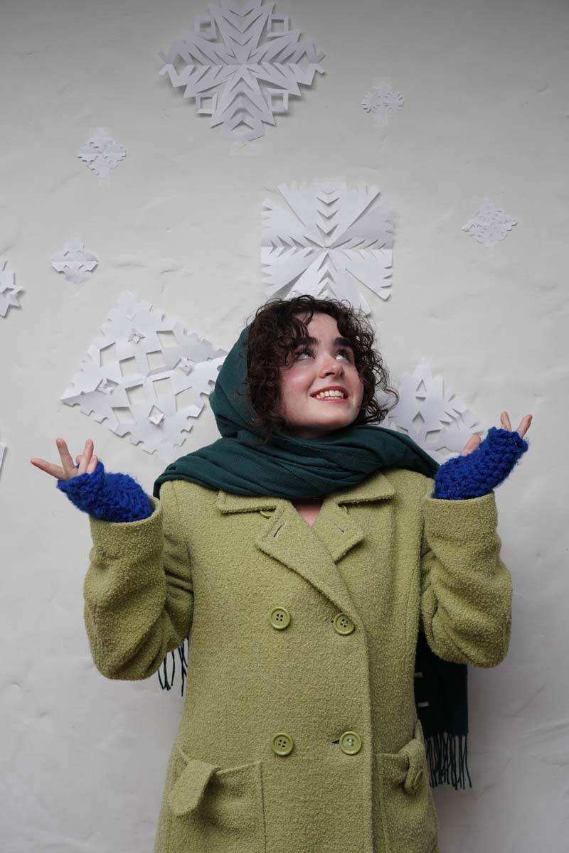 A person with long brown hair and a pea-green winter coat stands in front of some paper snowflakes, arms outstretched, smiling.