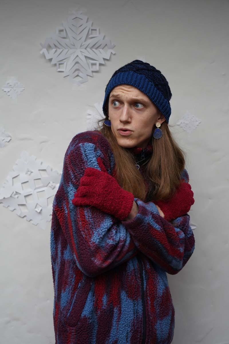 A person with long blond hair, a dark blue winter hat, and a jacket pretends to huddle while standing in front of some paper snowflakes.