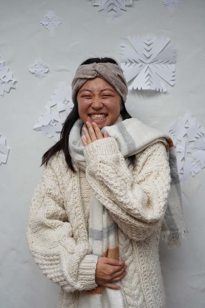 A person with long black hair wears a heavy cream-colored jacket while smiling and standing in front of some paper snowflakes.