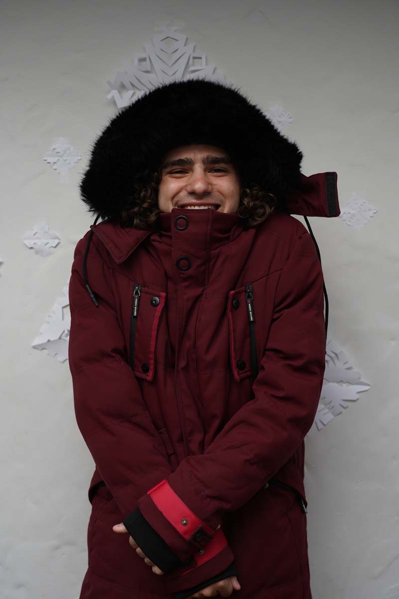 A person wearing a heavy black winter hat and a maroon coat stands in front of some paper snowflakes.