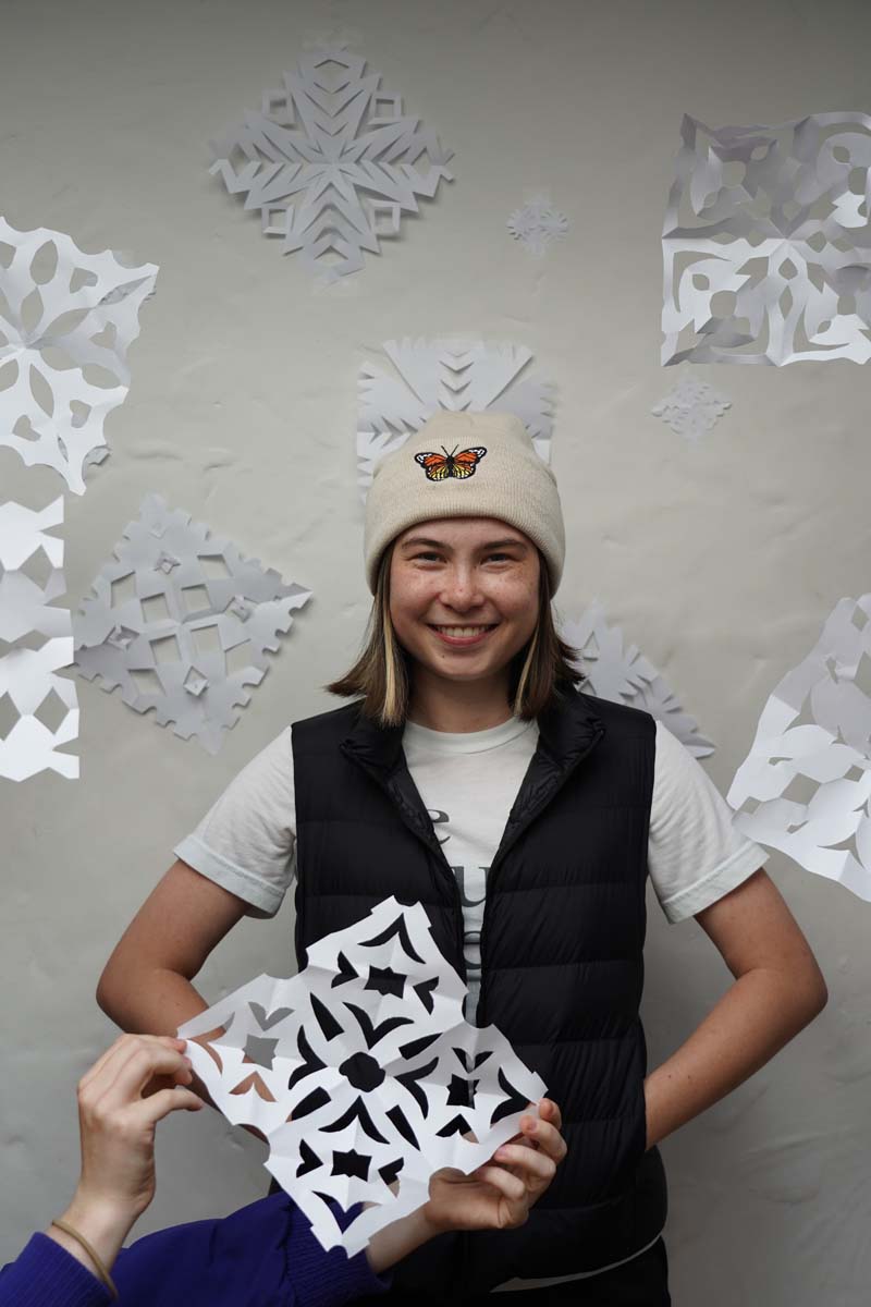 A person with long blond hair wears a cream-colored hat and a black vest. The person is holding a paper snowflake and standing in front of a wall of paper snowflakes.