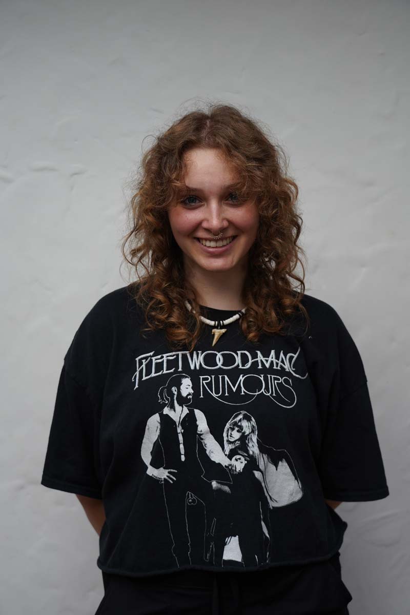 A person with long curly blond hair wears a black T shirt with the words "Fleetwood Mac: Rumors" on it. The person is smiling at the viewer.