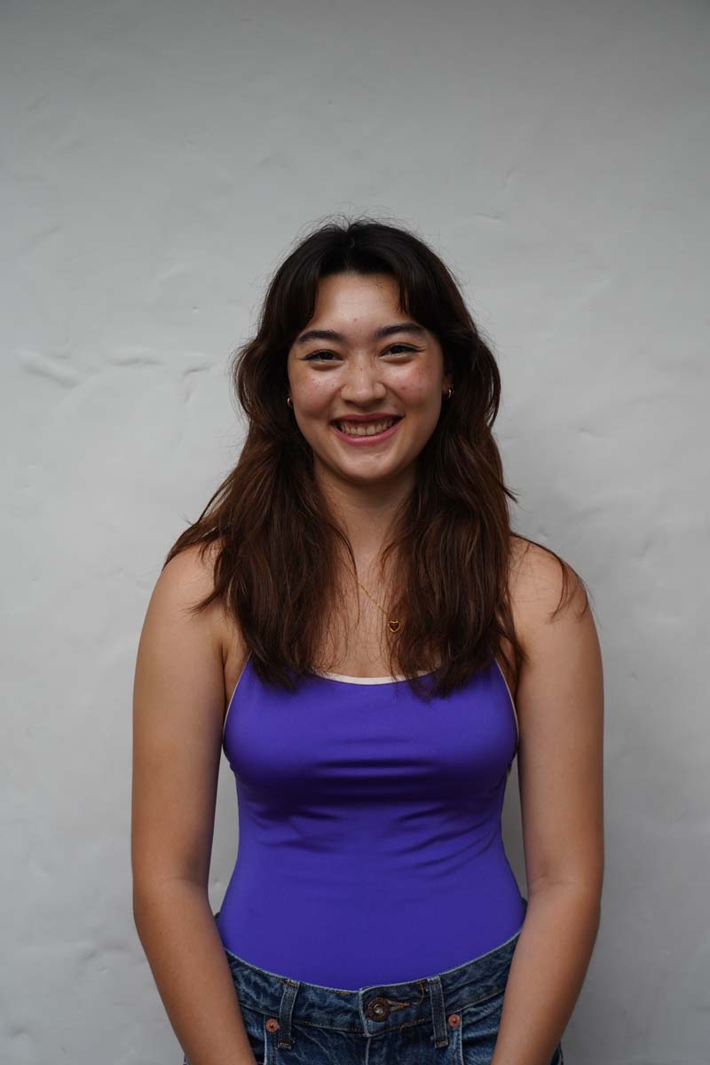 A person with long brown hair and a purple tank top smiles at the viewer.