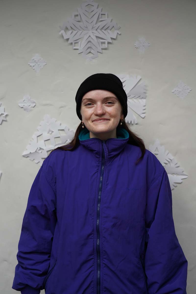 A person with a dark blue winter hair and a purple winter coat stands in front of a wall of paper snowflakes looking at the viewer.