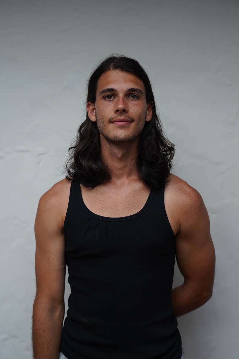 A person with long brown hair and a black tank top looks at the viewer.