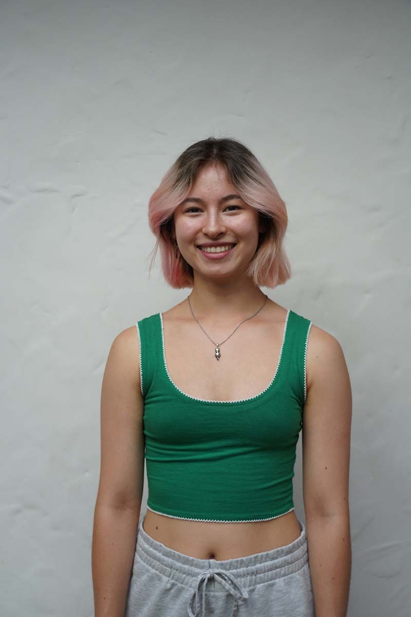 A person with shoulder-length pink-highlighted hair and a green tank top smiles at the viewer.