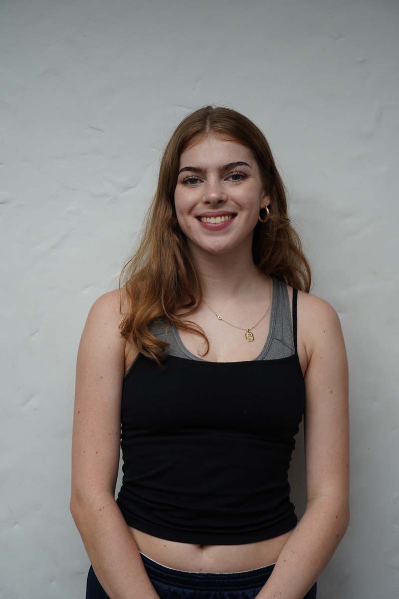 A person with long dark blond hair and a black tank top smiles at the viewer.