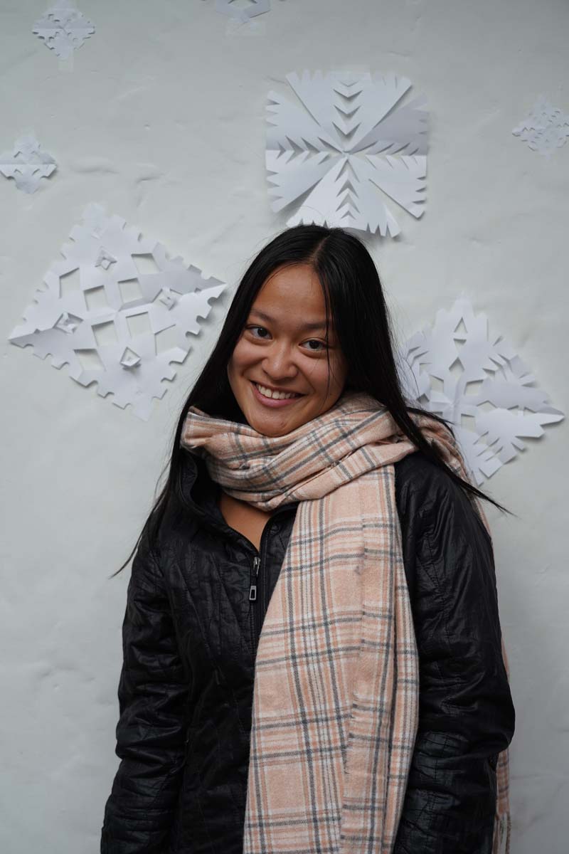 A person with long black hair and a black coat, wearing a heavy scarf, stands in front of a wall of paper snowflakes and smiles at the viewer.