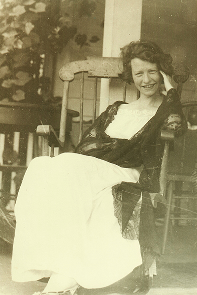 Edna St. Vincent Millay, smiling and wearing a long dress and lacy shawl, reclines in a rocking chair.