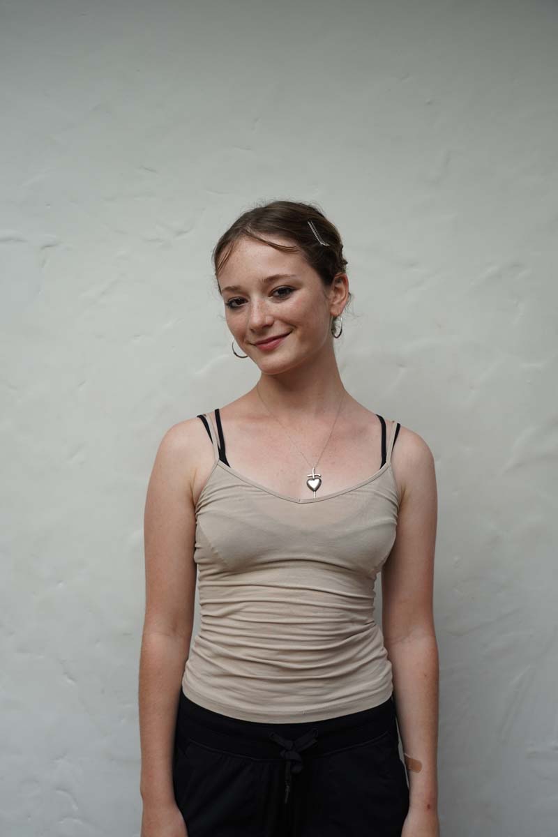 A person with long dark blond hair and a beige tank top smiles at the viewer.