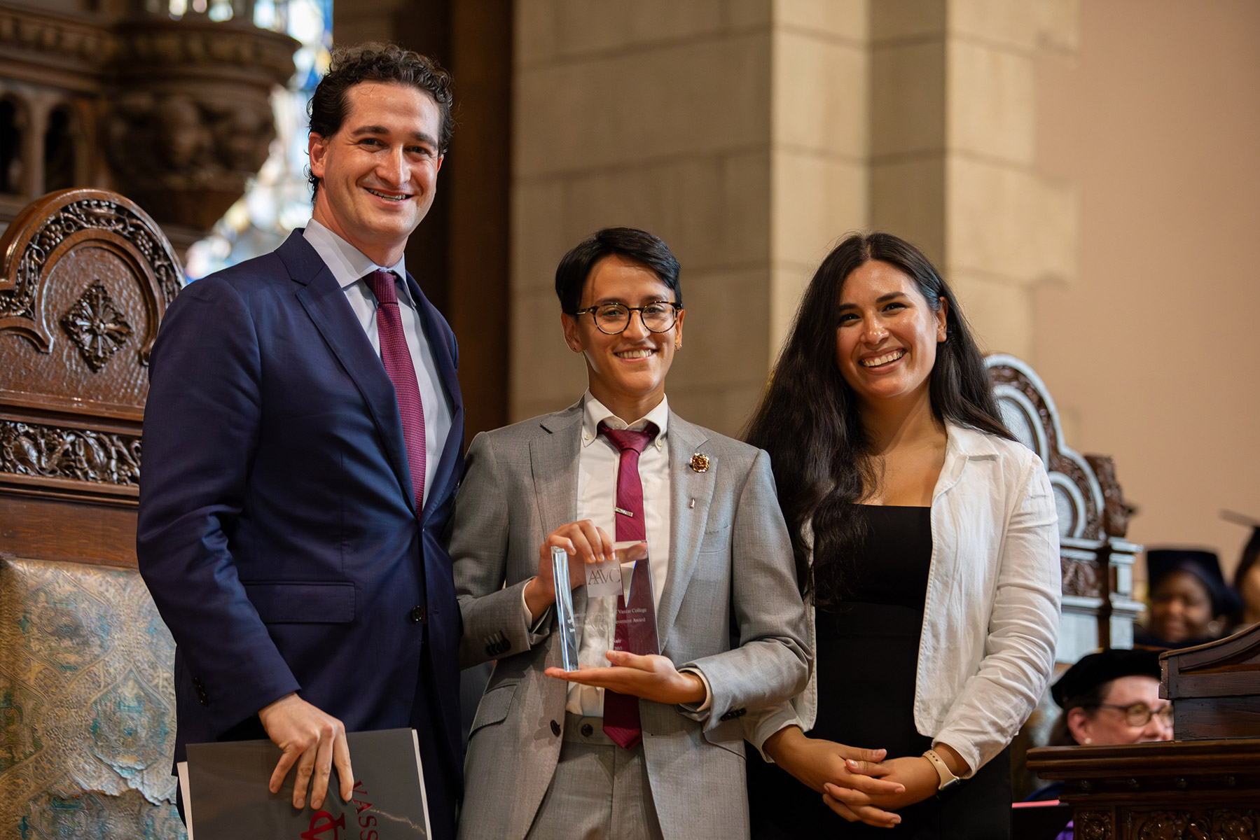 Three people standing and smiling on a stage with the person in the middle holding a glass award.