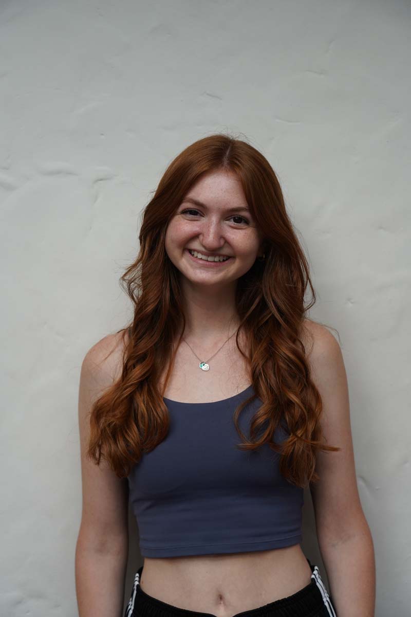 A person with long red hair and a dark blue tank top smiles at the viewer.
