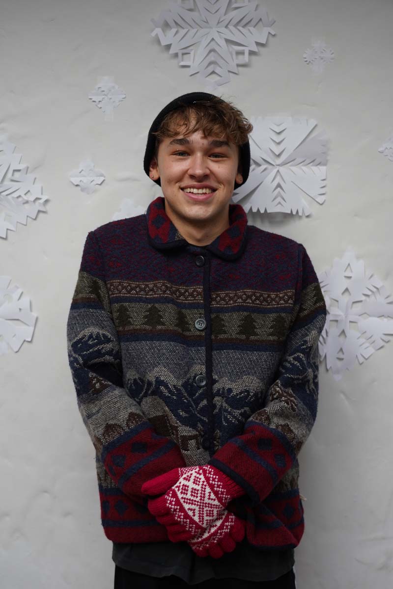 A person with short blond hair, a dark winter hat, and a patterned winter coat smiles at the viewer while standing in front of a wall of paper snowflakes.
