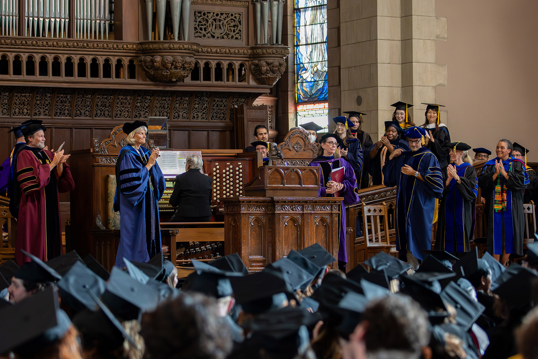 Person on stage surrounded by professors standing in formal graduation attire applauding with students in the front in caps and gowns applauding.