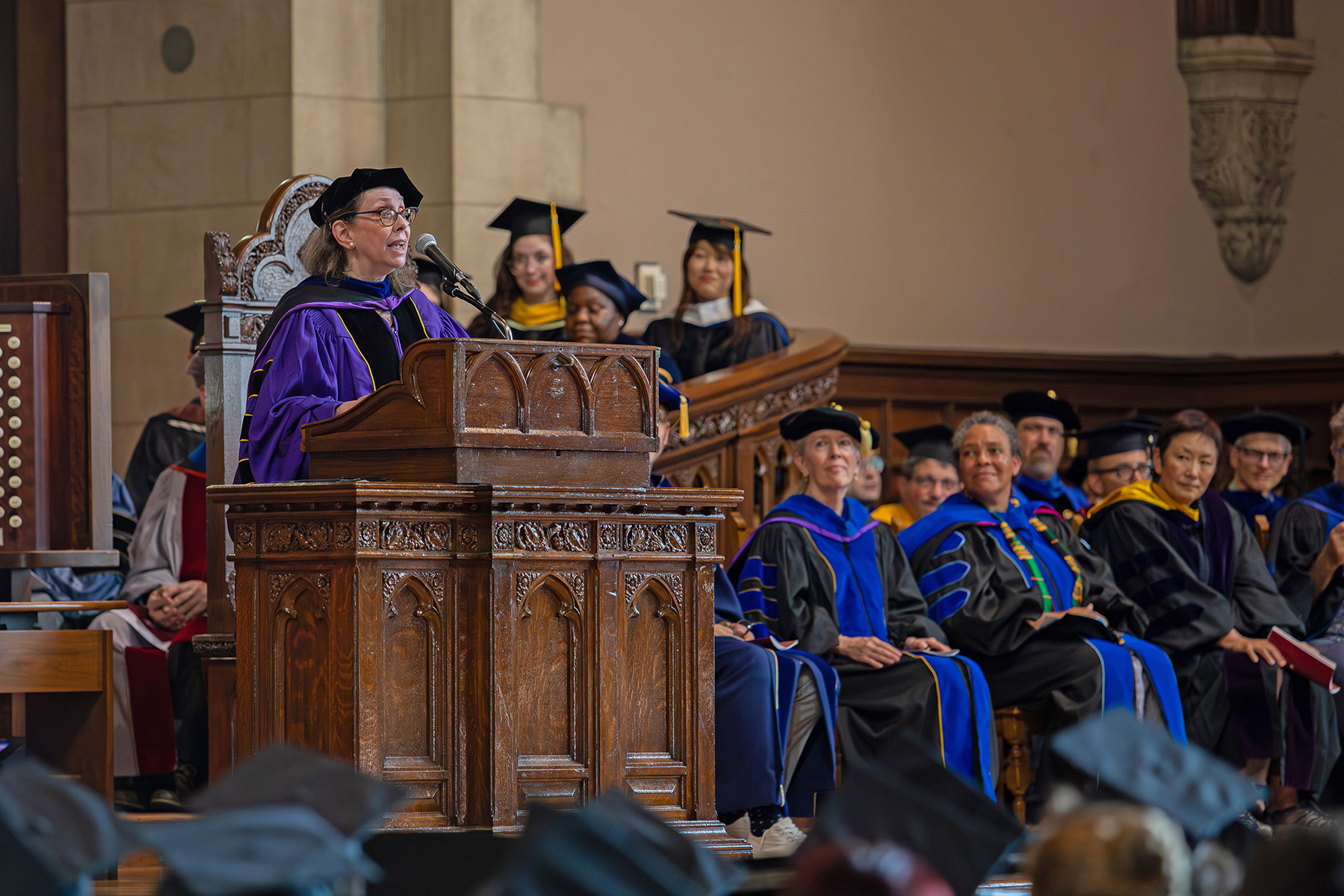 Person standing at a podium with professors sitting behind them and students in graduation caps sitting in front.