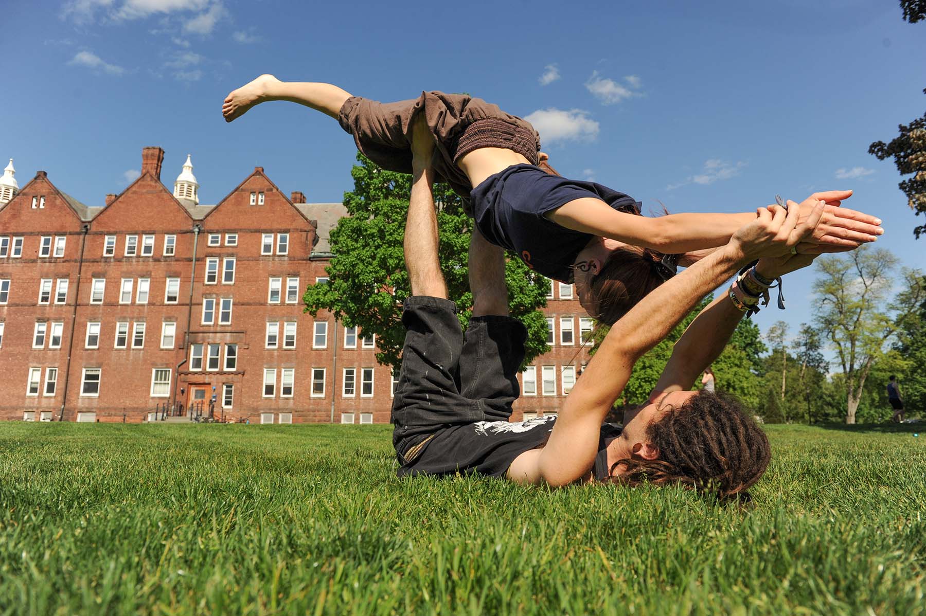 Two students practice yoga in the quad lying on the grass holding the other up in the air.