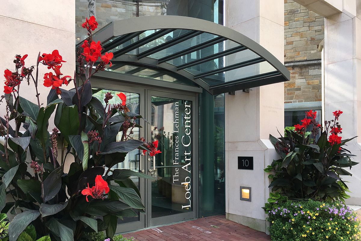 The front entrance of The Loeb featuring double glass doors, a curved awning made of metal and glass above, and flanked by tall red flowering plants.