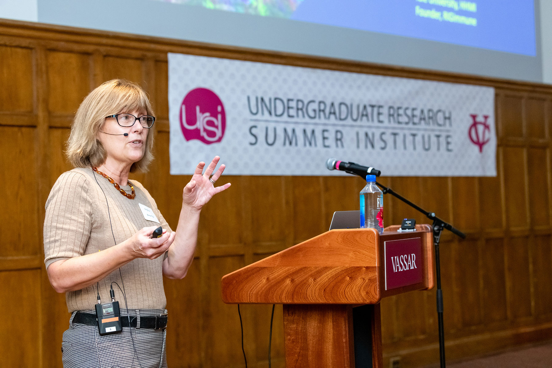 Person standing at a podium on a stage with a banner in the background that reads, "URSI: Undergraduate Research Summer Institute."