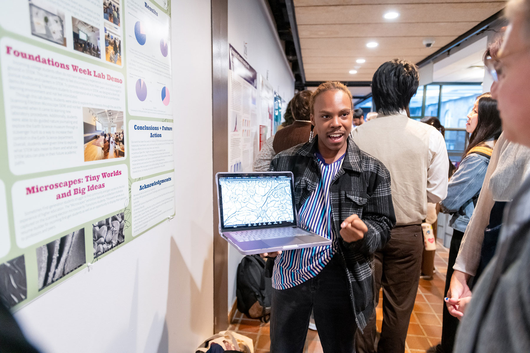 Person holding a laptop showing a person something on a screen with a poster on the wall is in the background.