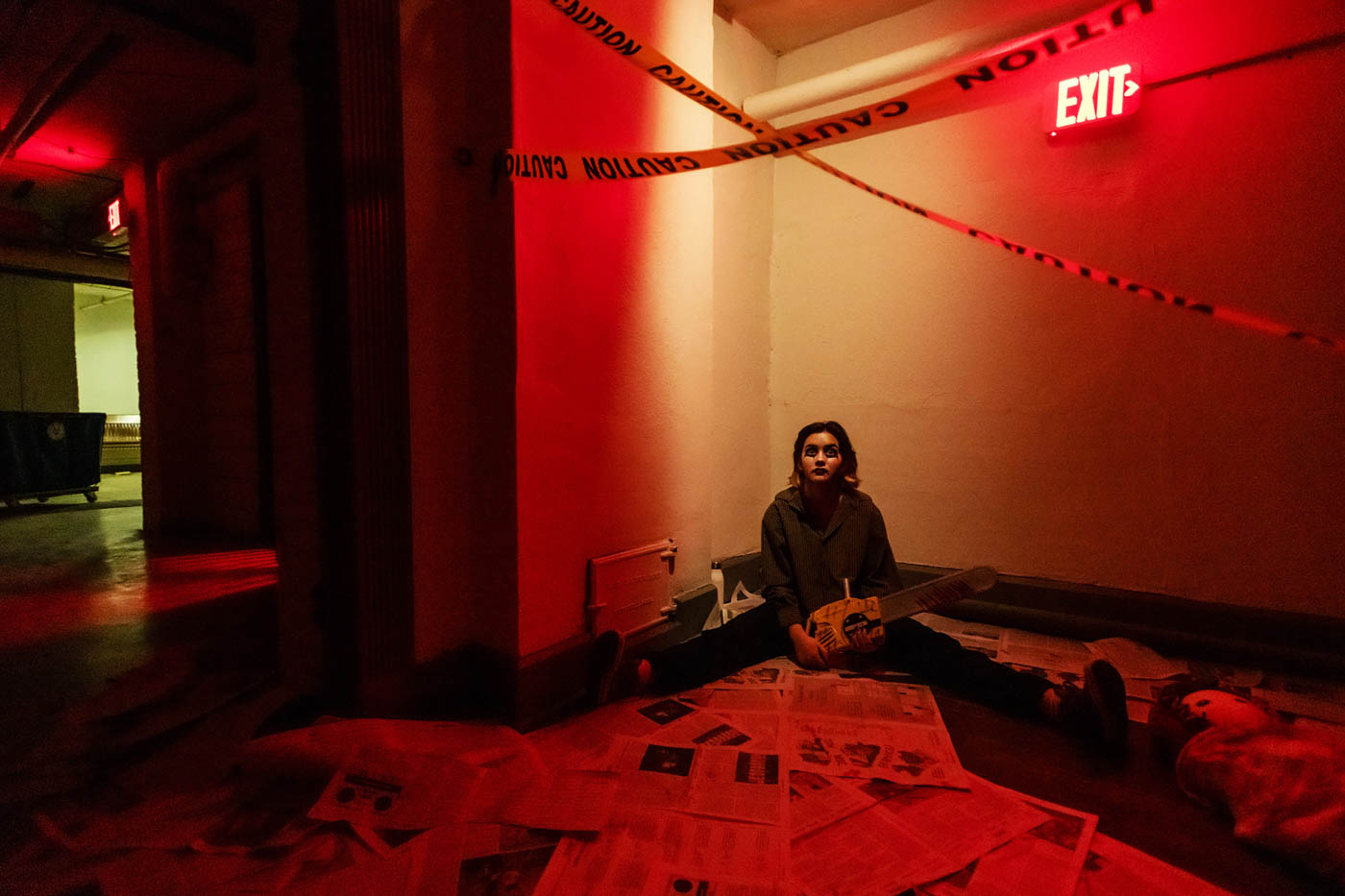 A haunted house. A person sits in a room, the walls lit only with red light, newspapers strewn around the floor. Yellow tape with “Caution” printed on it hangs from the ceiling. The person’s face is painted with black eyeliner, and the person is holding a chainsaw. This is a very creepy scene.