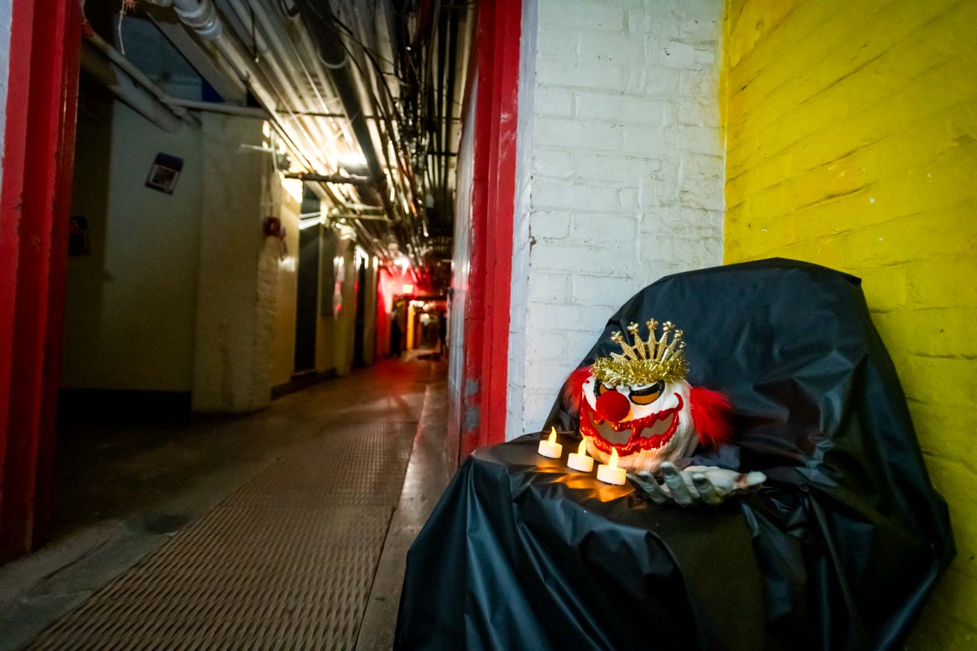 A long, narrow basement corridor, the ceiling crammed with pipes and cables. The foreground shows a chair draped in black, with three battery-powered candles on it. Also on the chair is a scary clown mask with a crown and gold wreath. No people are visible in this image.