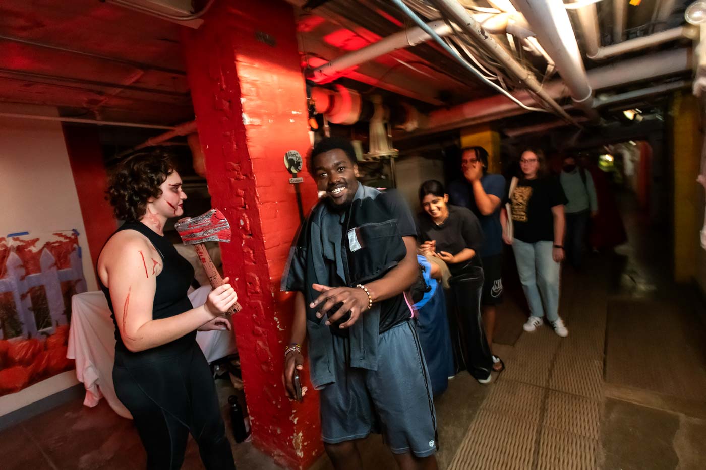 A basement hallway. Several people are standing in a line in the hallway. In the foreground is a brick pillar, painted bright red. Someone smeared with what is hopefully fake blood is brandishing a hatchet.
