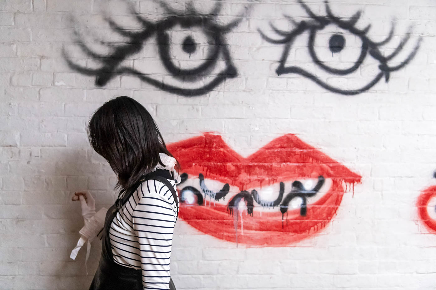 A person with long dark brown hair, a black dress, and a white-and-black-striped shirt stands next to a white brick wall on which is spray-painted a face with long eyelashes, bright red lips, and no nose.