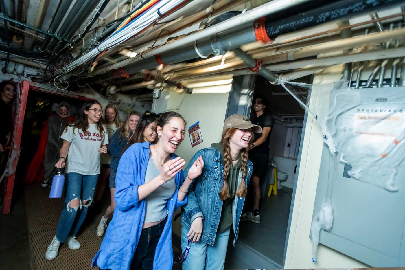 A group of people walks through a narrow hallway, the ceiling lined with thick arrays of pipes and cables. They look like they’re having a good time. In the background are several people with spooky face paint.