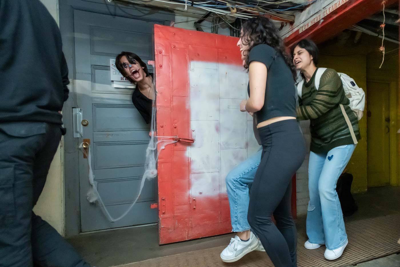 A person with long dark hair and heavy black facepaint around their eyes jumps out from behind a door, scaring people.