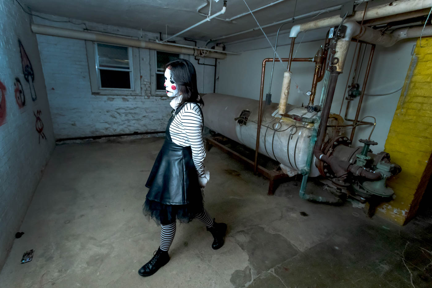 A large, ill-lit basement room with grimy brick walls, two small windows, and a large tank connected to various pipes. Pacing around this room is a person wearing a black dress, black-and-white-striped stockings and shirt, and black shoes. The person is wearing a white mask with bright red cheeks and lips and black holes for eyes. They look like a very creepy rag doll.