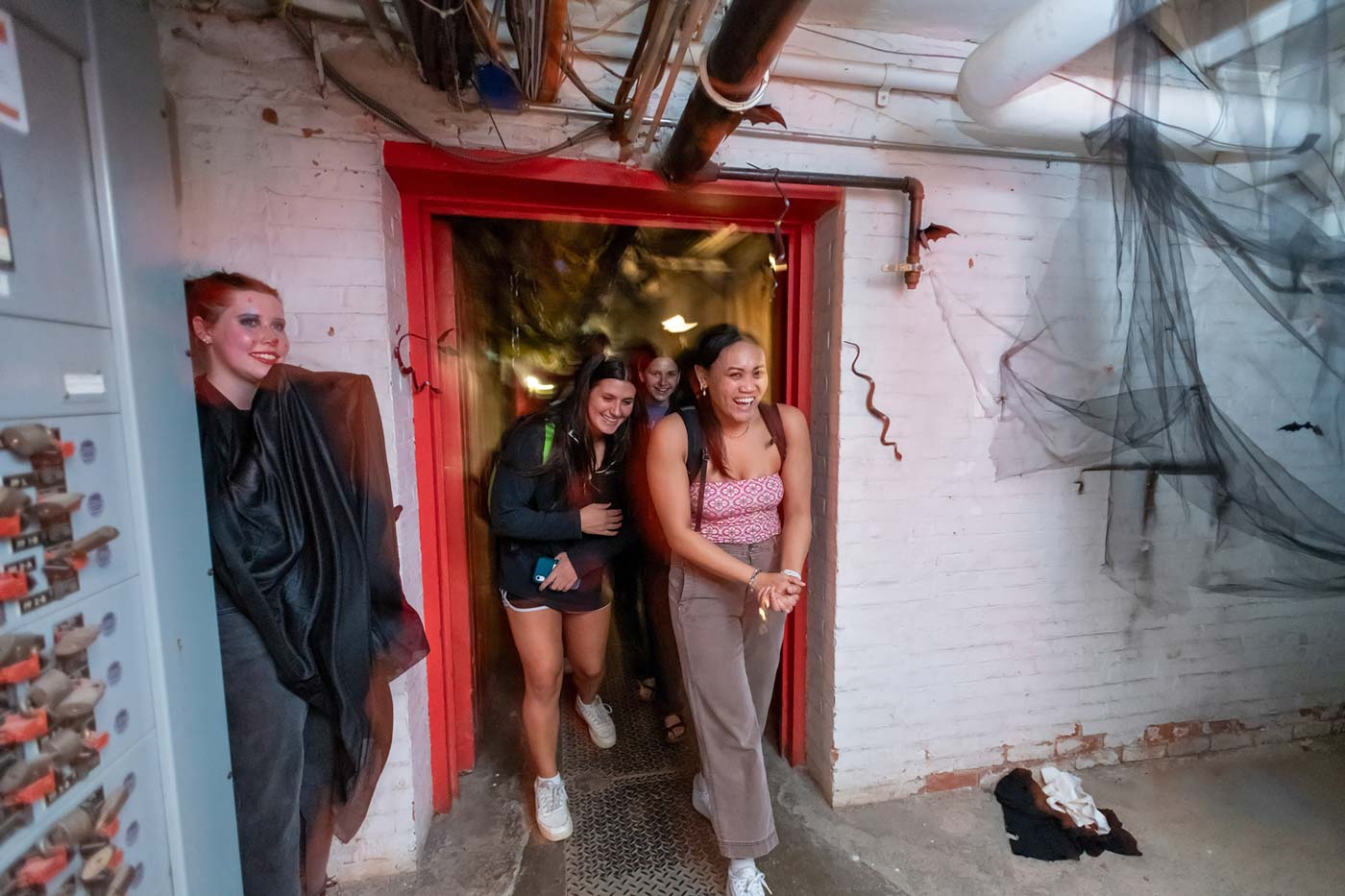 People walk through a red-framed doorway into a dingy room with white brick walls and black netting, probably meant to represent spiderwebs, hung from the ceiling.