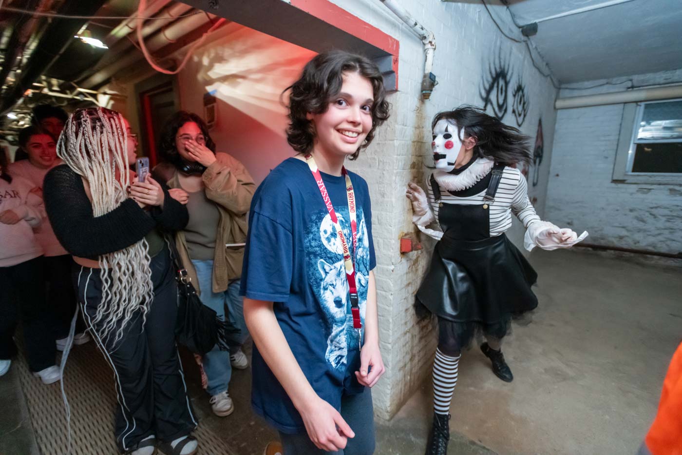A person in a black dress and black-and-white-striped stockings and shirt jumps out at some people from behind a wall. The person is wearing a white mask with bright red cheeks and lips and black holes for eyes. Someone stands in the foreground with long brown hair and a blue shirt with a wolf illustration on it, smiling at the viewer.