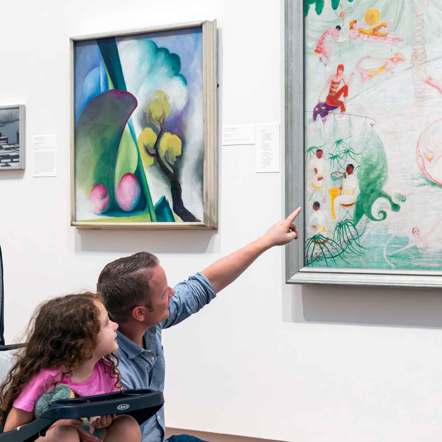 Adult and child crouching and below a painting on the wall while looking at. The adult is pointing at the painting.