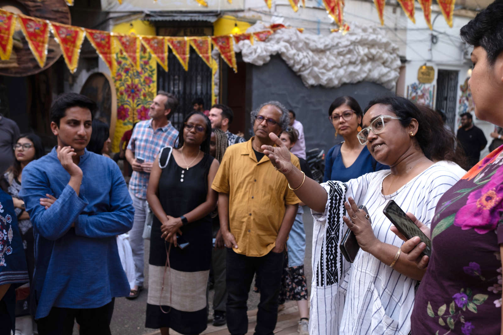 A group of people stand in a crowded street. A person with long black hair, glasses, and a white striped dress is pointing to something off camera.