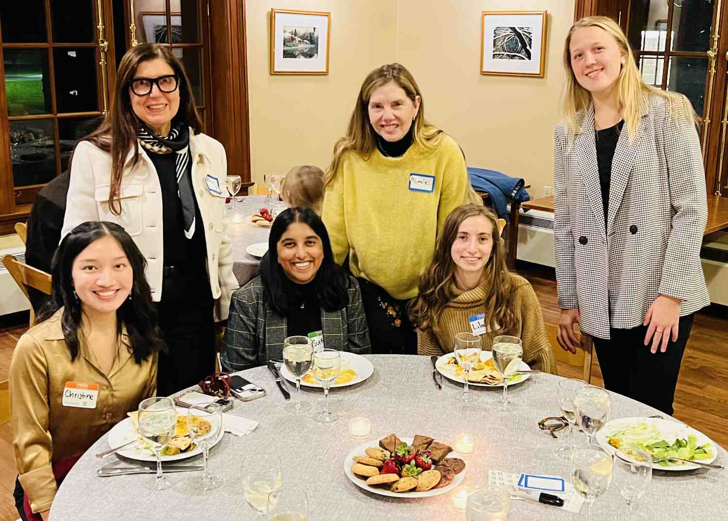 Six people are in a room with a hardwood floor and pictures on the walls. Three are sitting at a round kitchen table with food on it. All are smiling at the camera.