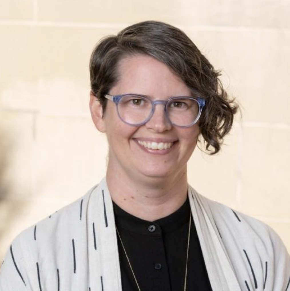 A person with brown hair, short on the right side, and long on the left; purple-rimmed glasses; and a white shirt smiles at the viewer.