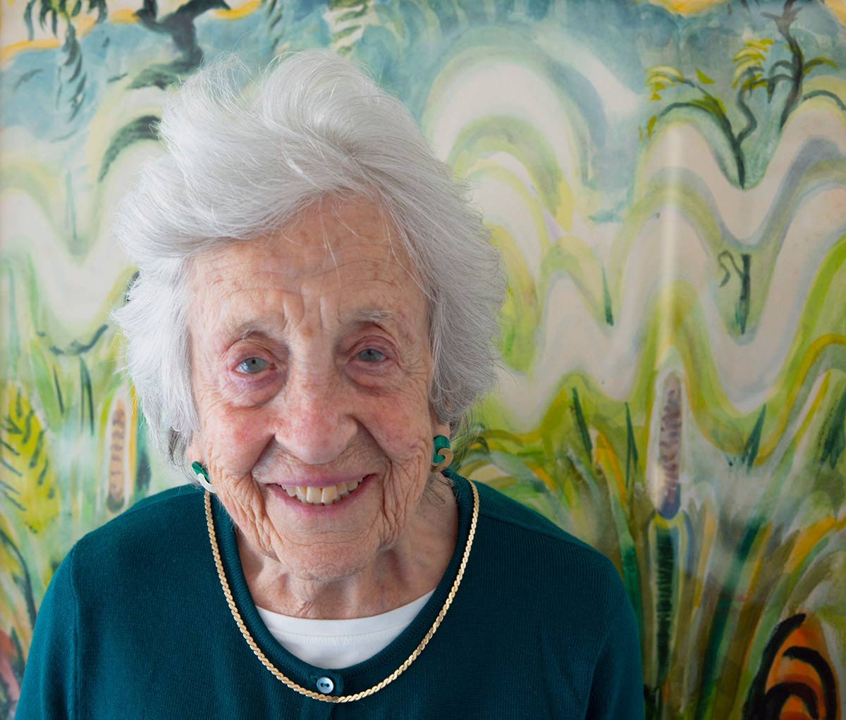 A person with mid-length gray hair, a dark blue sweater, and a gold necklace sits in front of a painting of plants, smiling at the viewer.