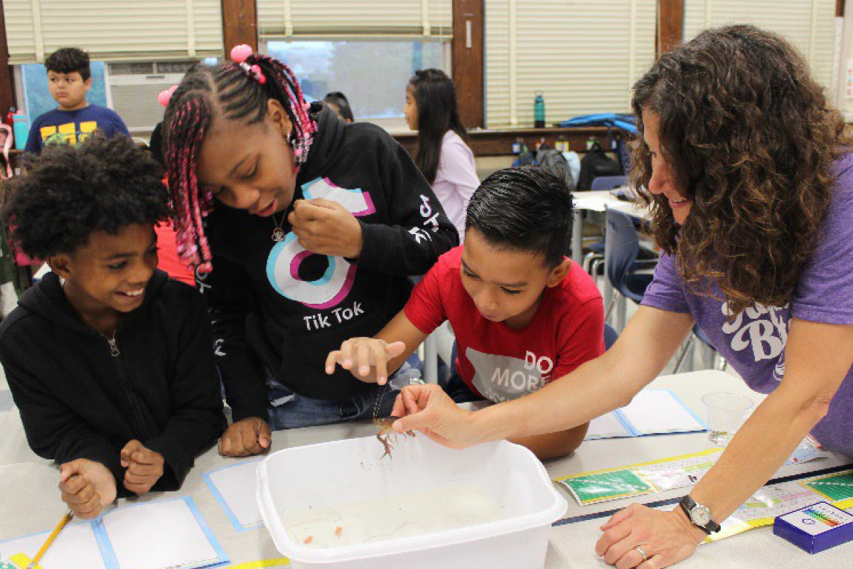 Jennifer Rubbo shows a crayfish to three elementary school students as they all gather around a table.