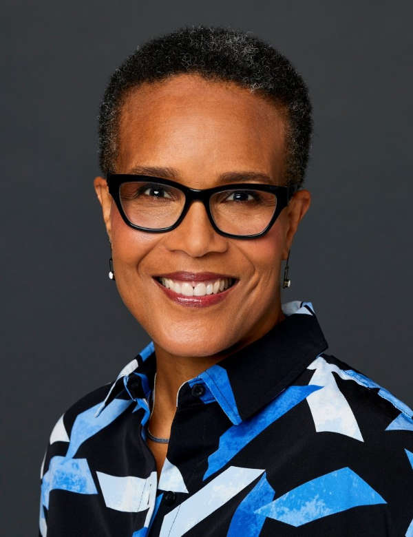 A person with short black curly hair, black-rimmed glasses, and a brightly patterned black and blue shirt smiles at the viewer.