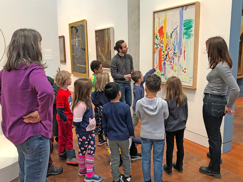 A guide with a group of children and a teacher view a painting in a gallery.