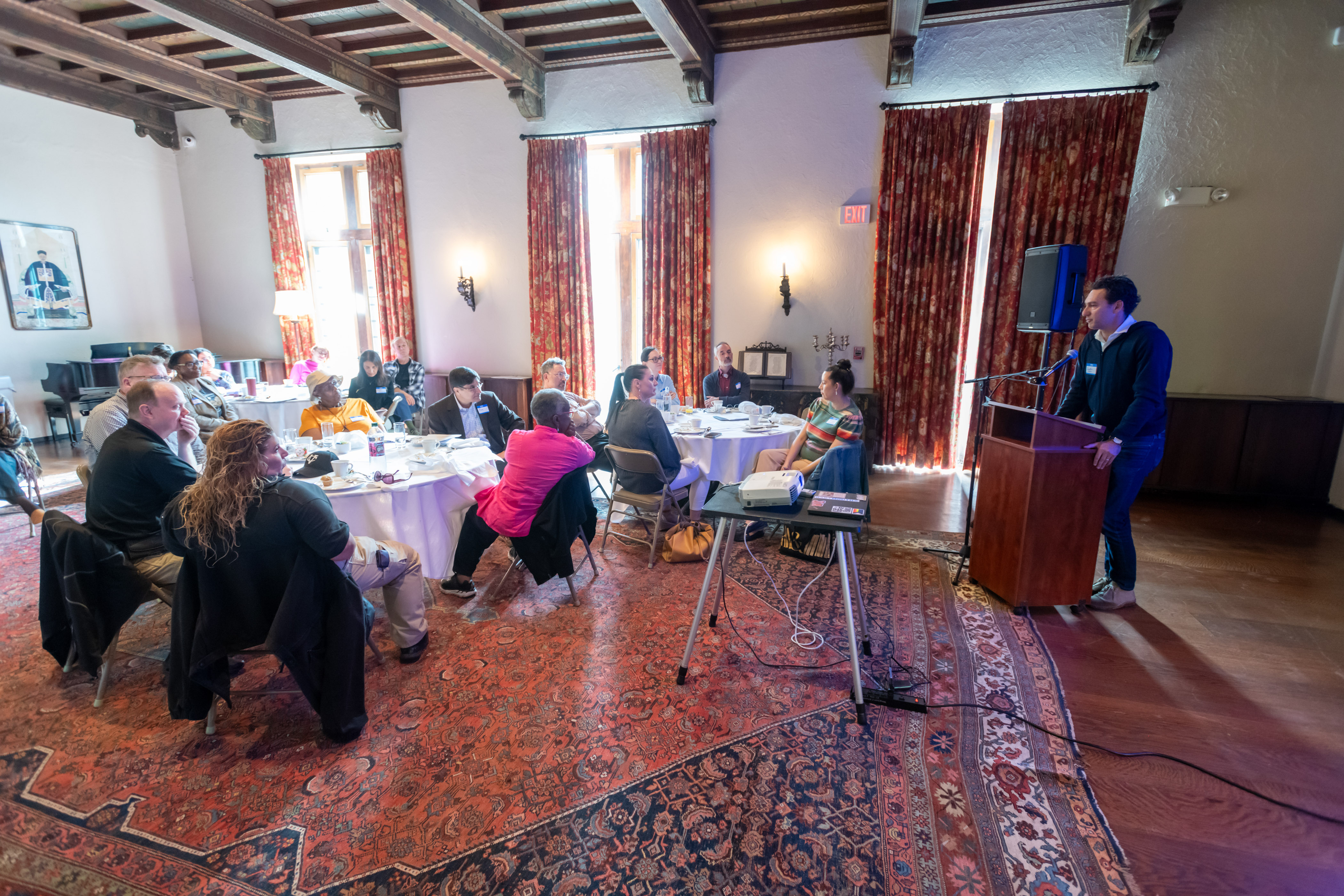 A large, very fancy room with a wood-paneled ceiling, tall windows, and several ornate rugs. There are several round tables in the room, at which people are sitting. On the right side of the photo is a person standing at a wooden podium, speaking to the group.