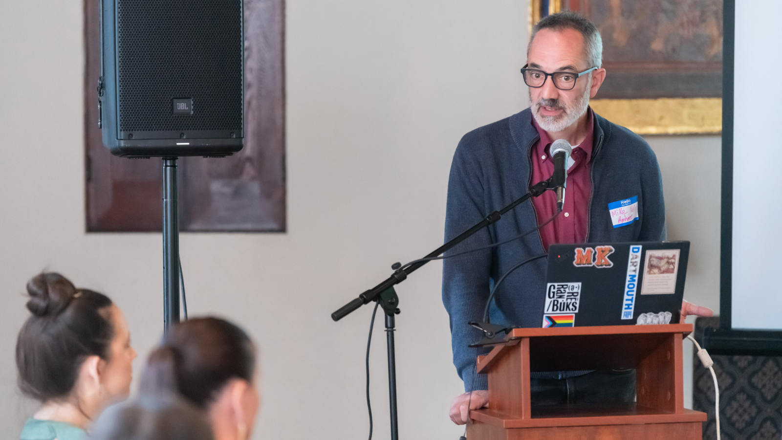 A person with short gray hair, a gray beard, and black-rimmed glasses speaks at a microphone in a room with oil paintings on the wall. The person is standing in front of a laptop with stickers on the lid. Several people are listening. 