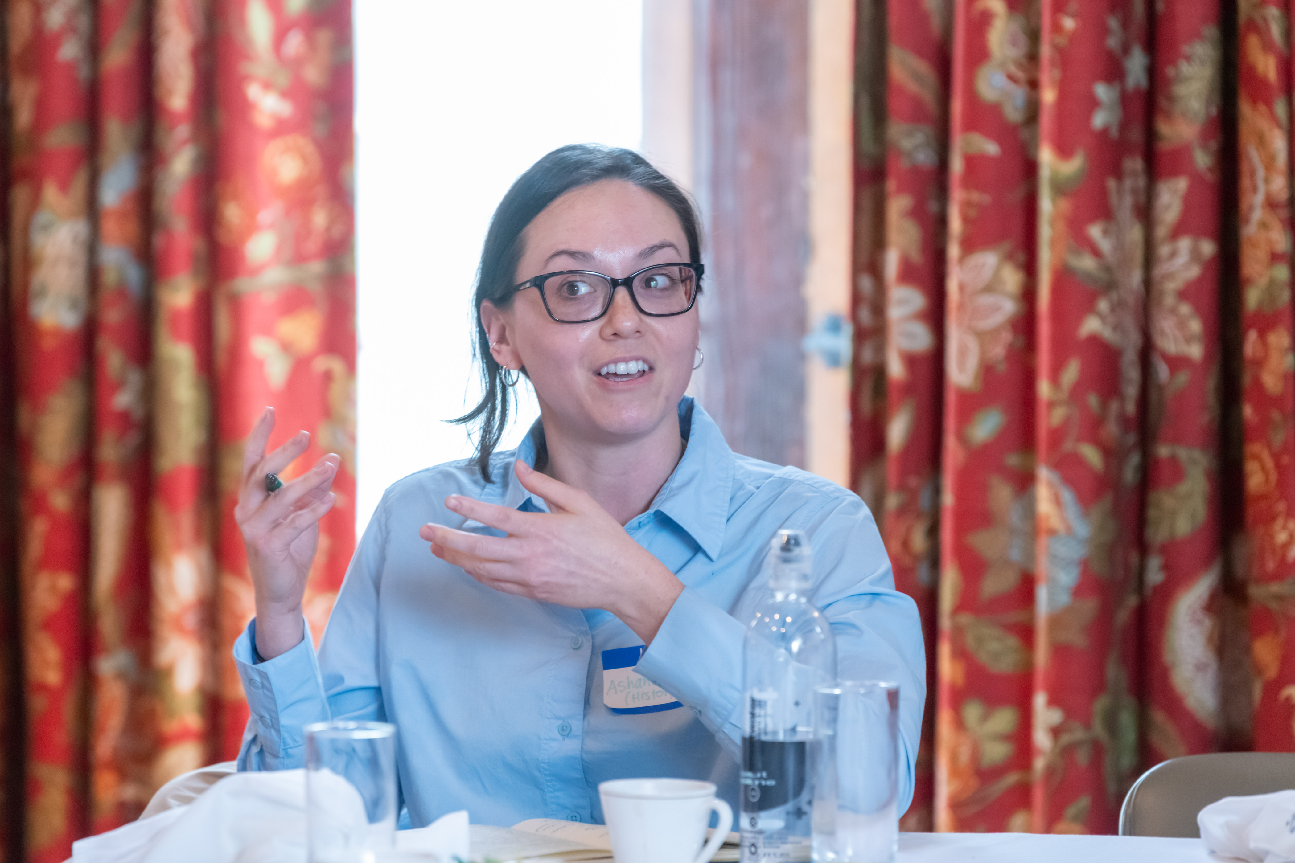 A person with long dark hair, black-rimmed glasses, and a blue button-down shirt sits at a table with glasses on it. The person is speaking to others not shown in the photo.