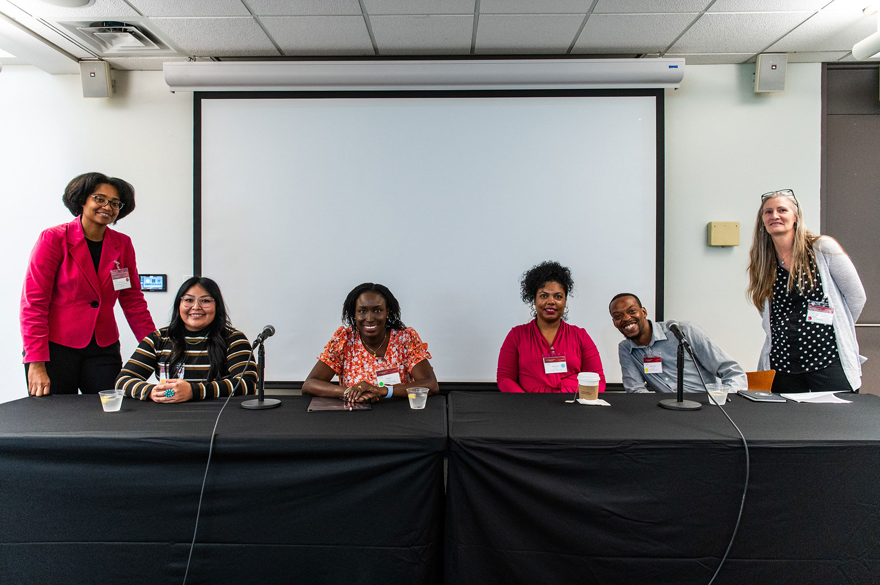 Six smiling people sitting at a covered table with a projector screen in the background.