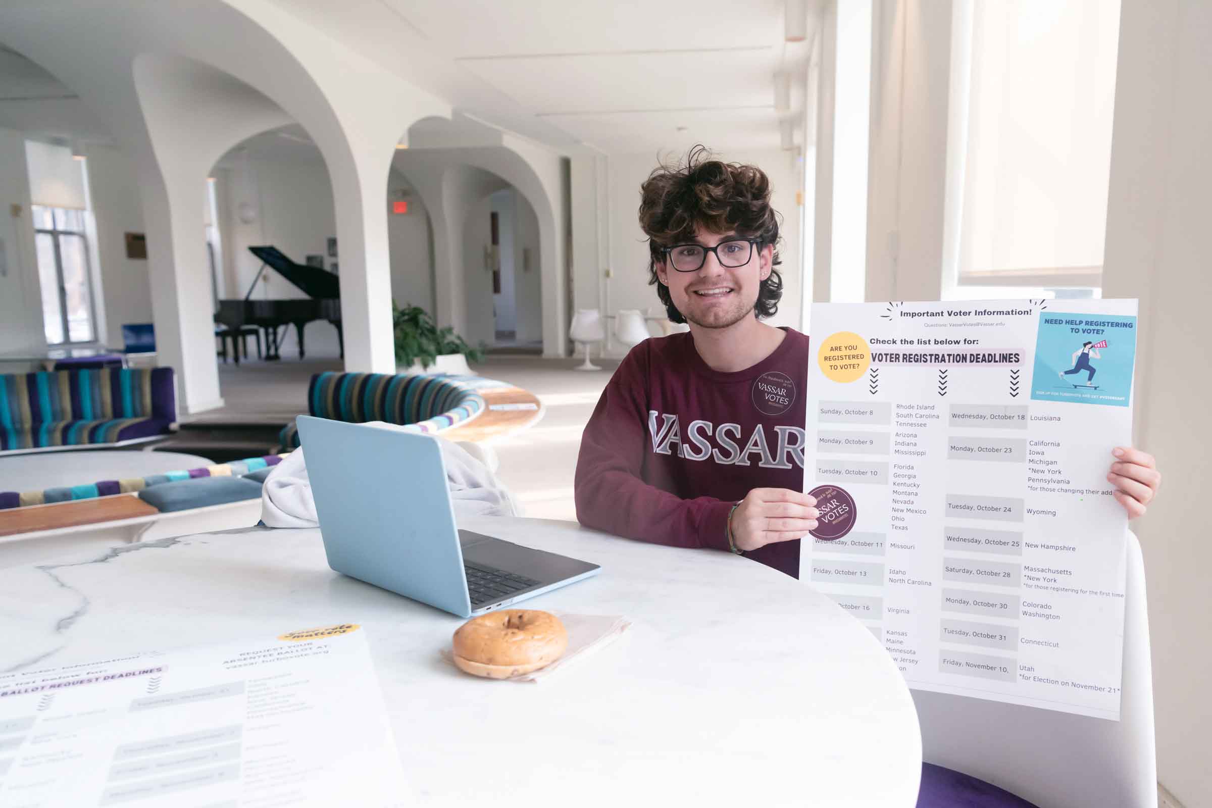A smiling person sits at a table with a bagel and a laptop holding up a voter registration deadline poster.