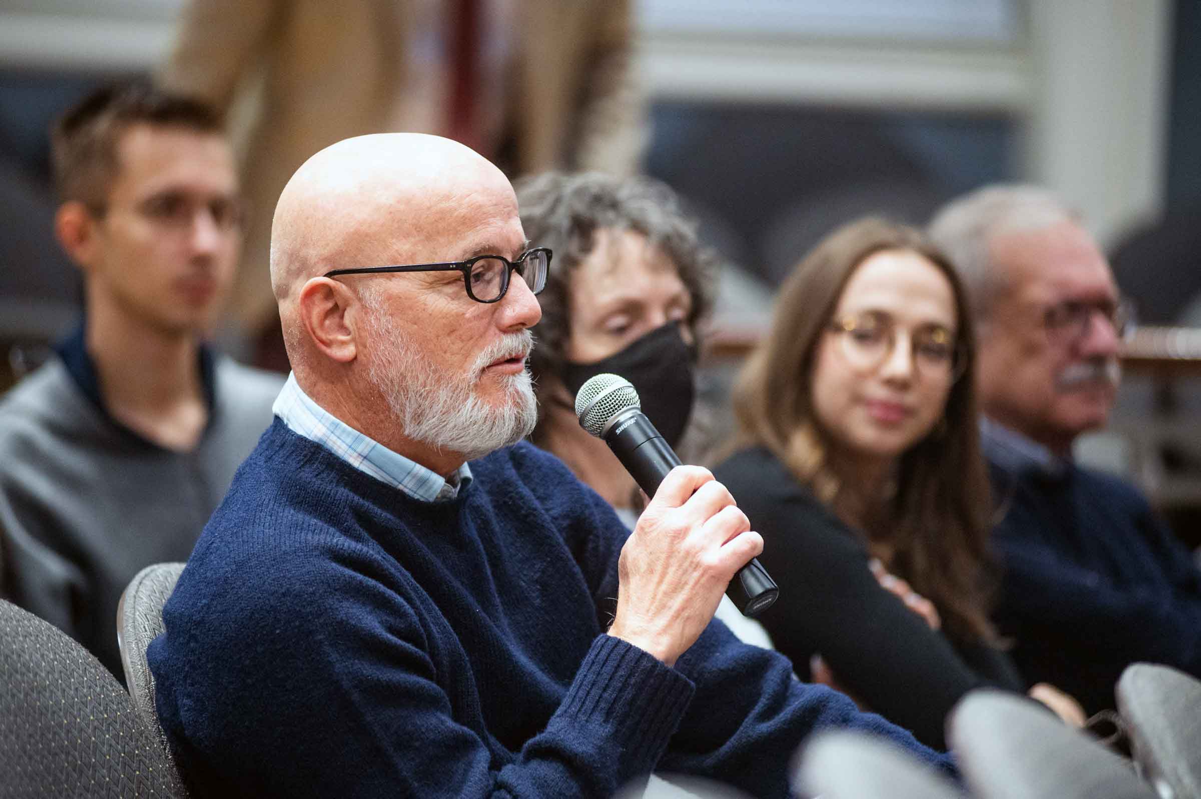 Seated person with a glasses and a beard speaking into a microphone while others in the background watch and listen.